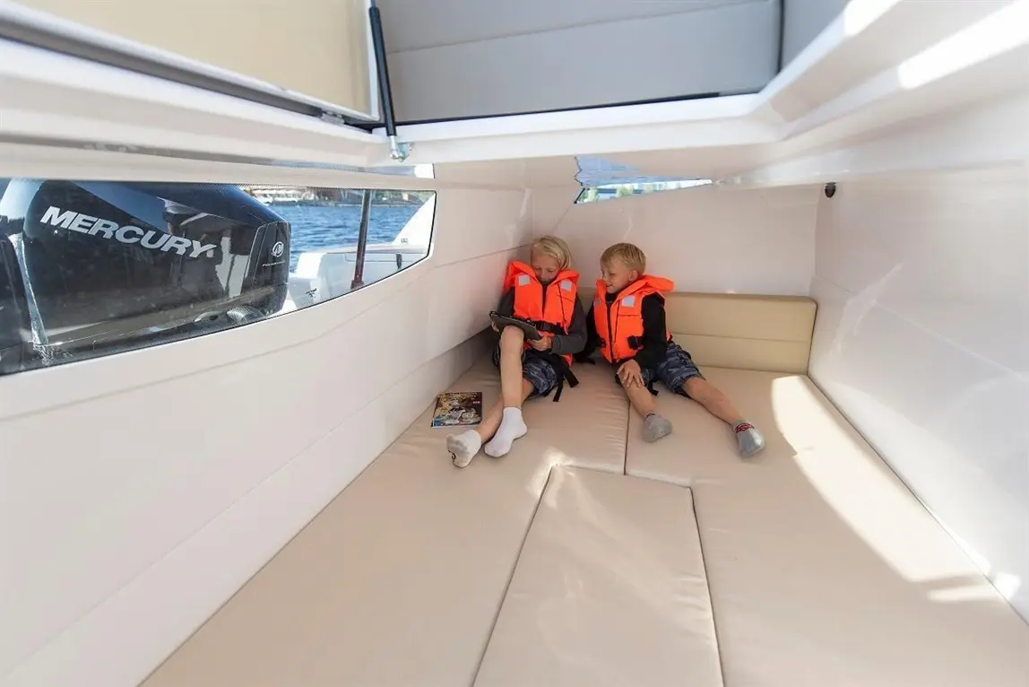 Two children in bright orange life jackets sit on a cushioned boat interior, using a tablet while a Mercury outboard motor is visible.
